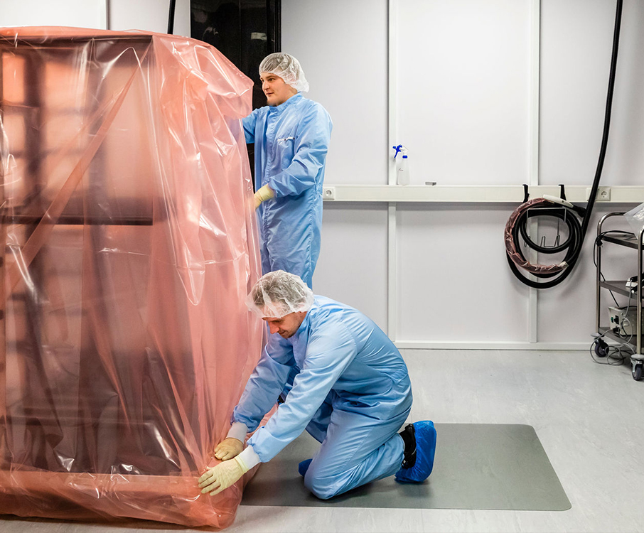 Twee medewerkers van Faes voeren een high-tech verpakkingsproces uit in een cleanroom, waarbij een grote component zorgvuldig wordt ingepakt.