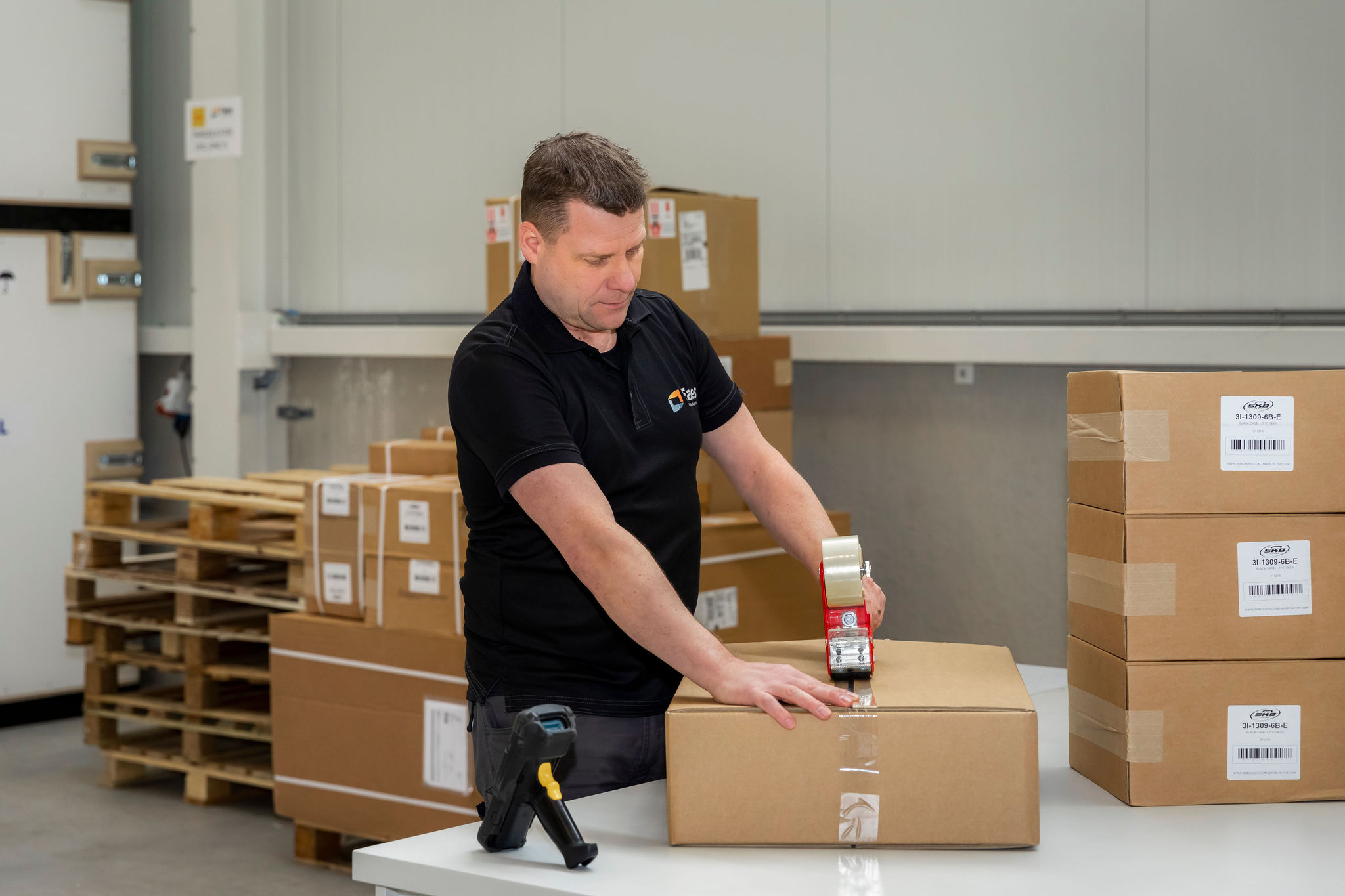 Een Faes-medewerker sluit zorgvuldig een kartonnen doos met een tape-dispenser in een magazijn, omringd door andere stapels kartonnen dozen. De medewerker heeft ook een barcode scanner bij de hand voor nauwkeurige tracking.