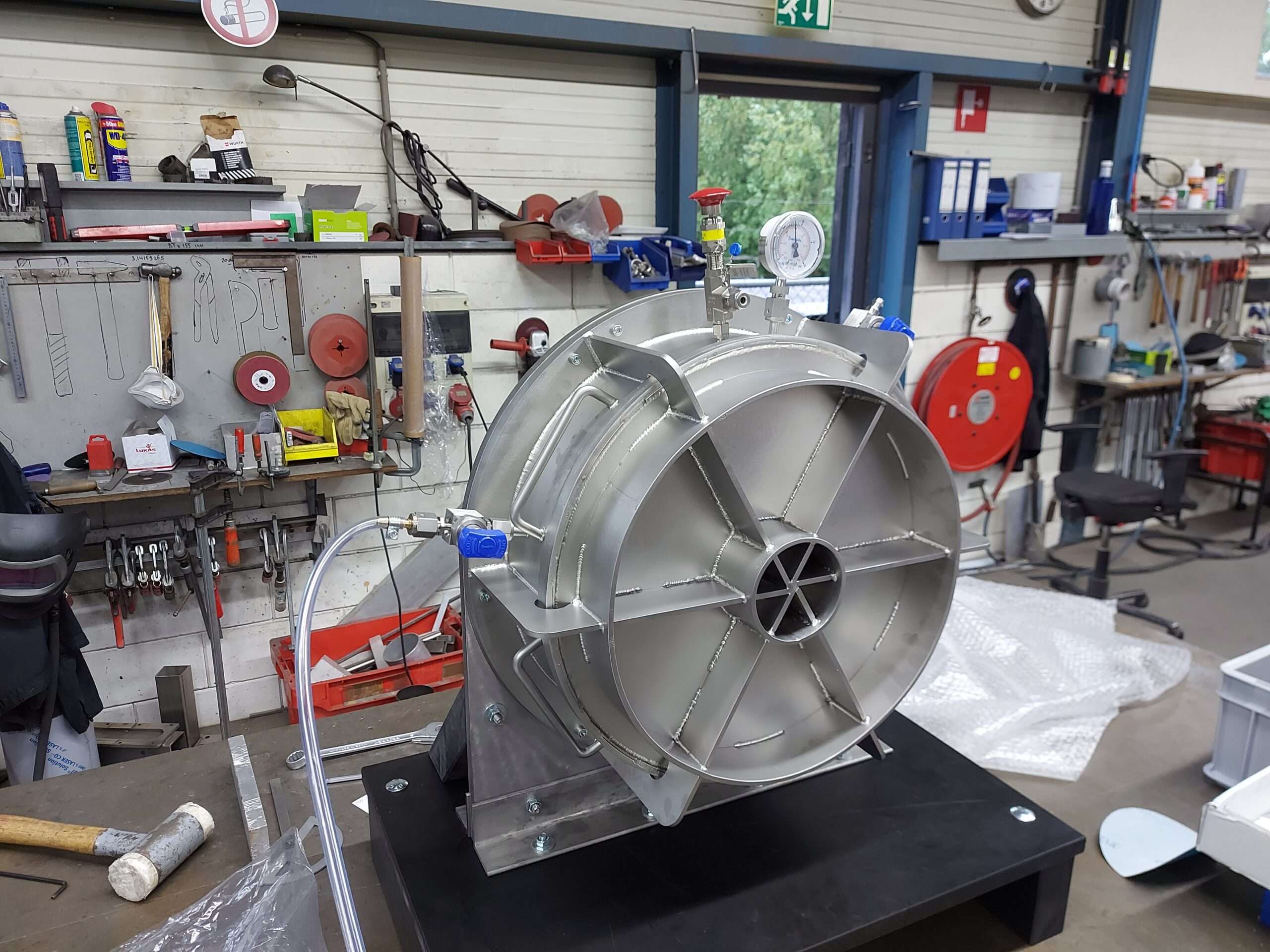 Een roestvrijstalen hoogdruk systeem in een werkplaatsomgeving, uitgerust met slangen, manometers en kleppen. Dit apparaat is ontworpen voor geavanceerde toepassingen in de hightech industrie, met precisiecomponenten voor drukregeling en veiligheid. Het systeem is nog in de montagefase, omringd door gereedschap en werkbankaccessoires.