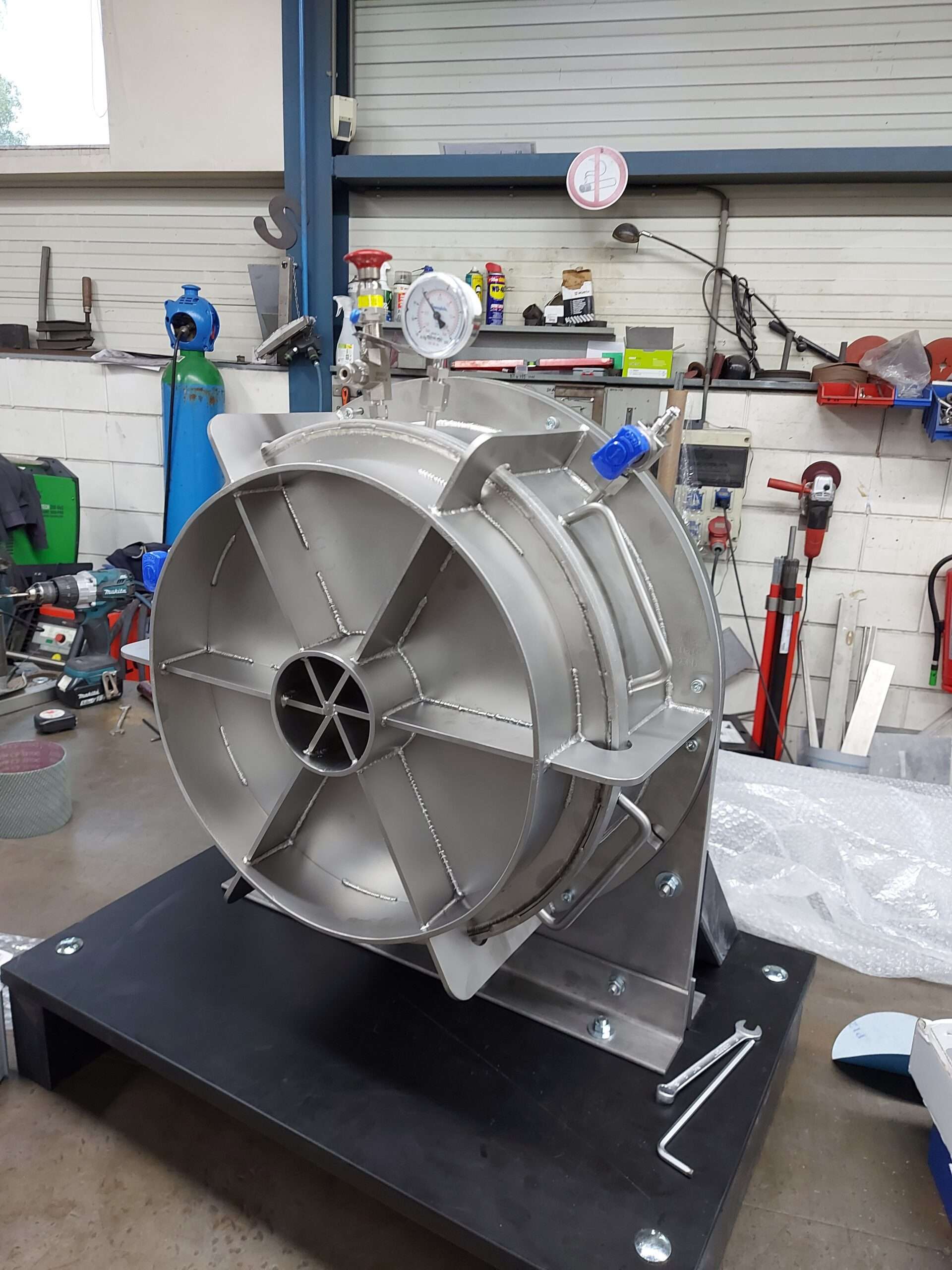 Roestvrijstalen dome geïnstalleerd op een stevige basis in een industriële werkplaats. De dome is uitgerust met diverse aansluitingen en meetinstrumenten, waaronder een manometer. De omgeving toont een georganiseerde werkruimte met gereedschappen, gasflessen en andere assemblage-uitrustingen, wat wijst op een technisch complexe setting voor het bewerken en assembleren van hightech componenten.