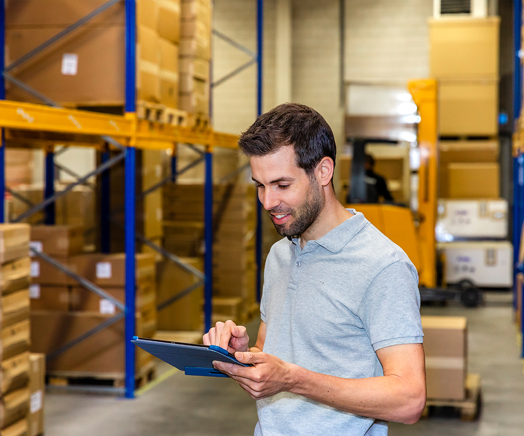 Een magazijnmedewerker gebruikt een tablet voor verpakkingsbeheer in een opslagruimte met stapelrekken en een heftruck op de achtergrond.