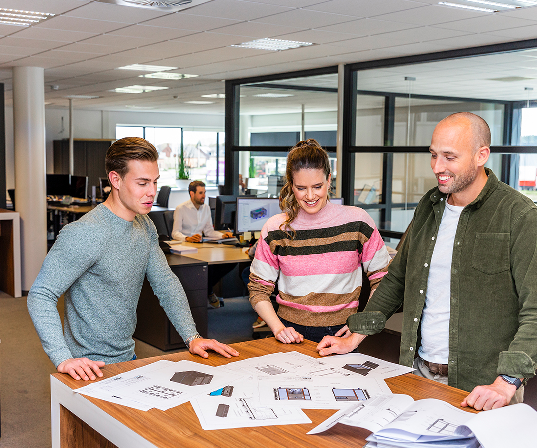 Een team van Faes medewerkers bespreekt verpakkingsontwerpen aan een tafel, met verschillende technische tekeningen en blauwdrukken verspreid.